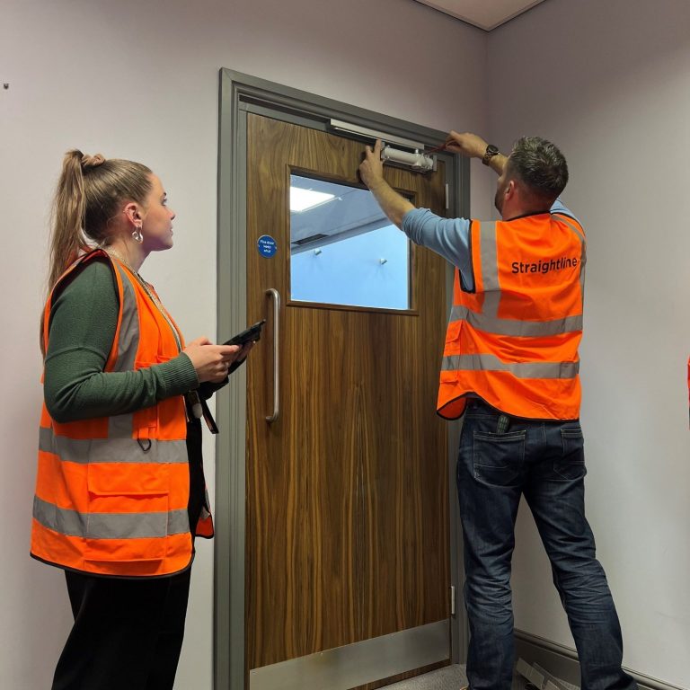 Entretien de porte coupe-feu Deux personnes en gilets orange inspectent une porte près d'un mur intérieur.