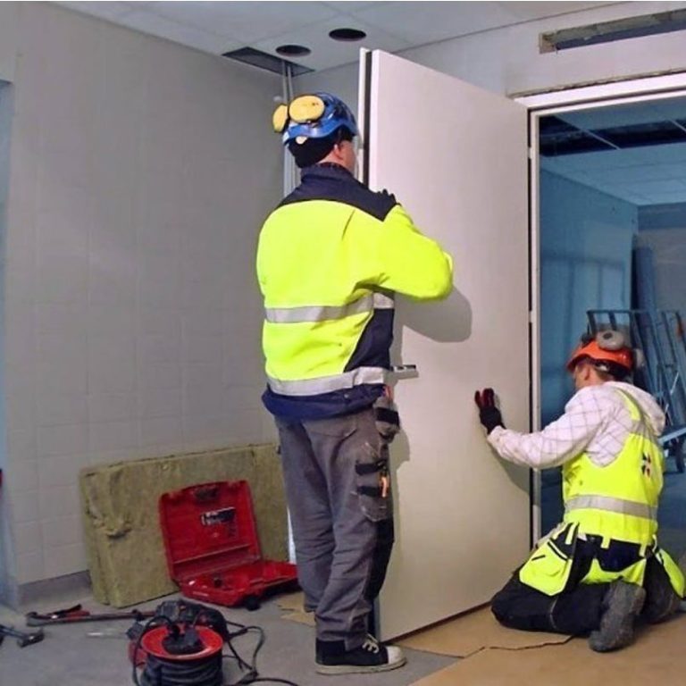 Installation de porte coupe-feu Deux travailleurs en tenue de sécurité installent une porte dans un bâtiment.